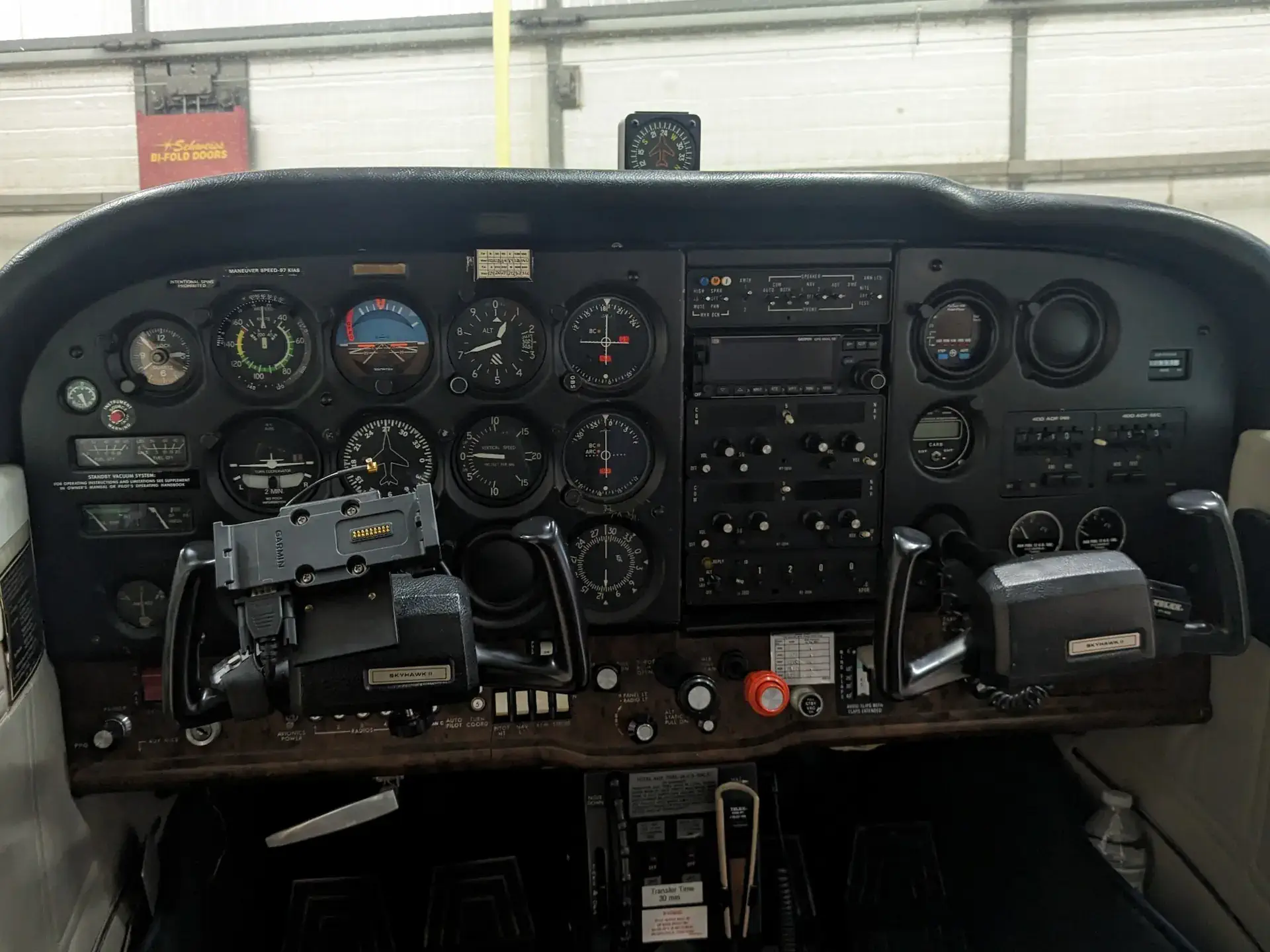 Cockpit instrument panel of a small aircraft showing various gauges, switches, dials, and two control yokes inside a hangar.